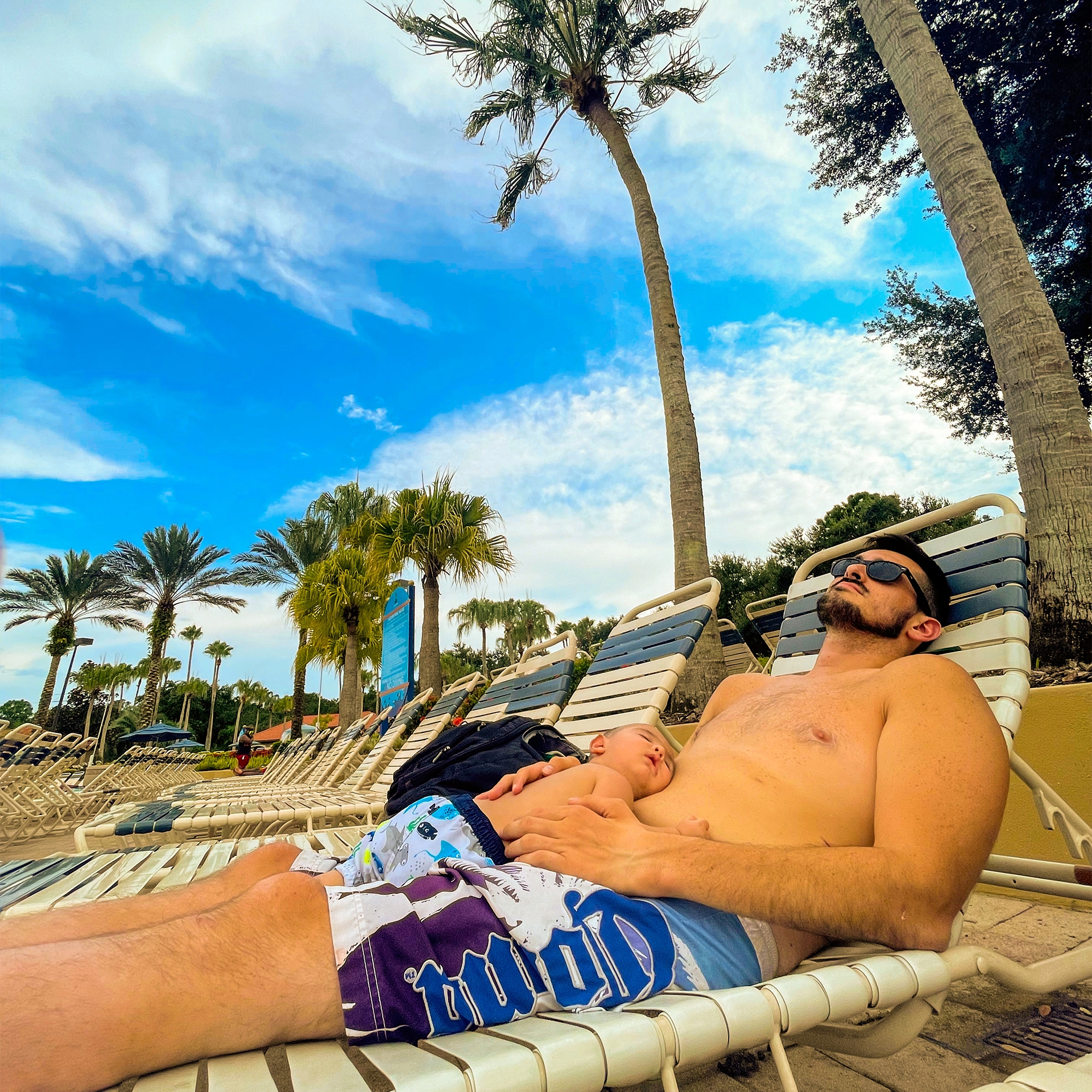 A man and an infant lay on a beach lounge chair under palm trees.