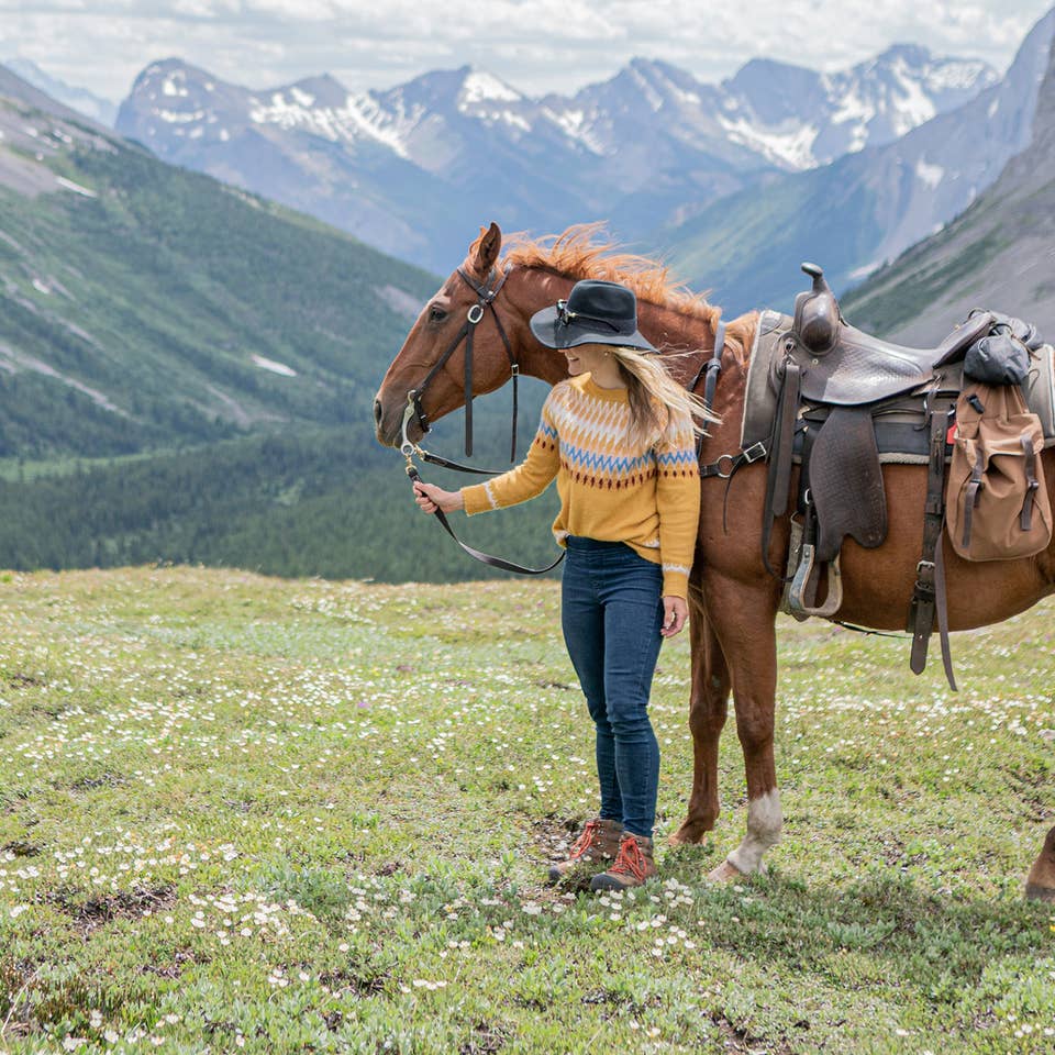 Featured Contributor, Ashlyn George, stands with a horse in front of the Canadian mountain range.