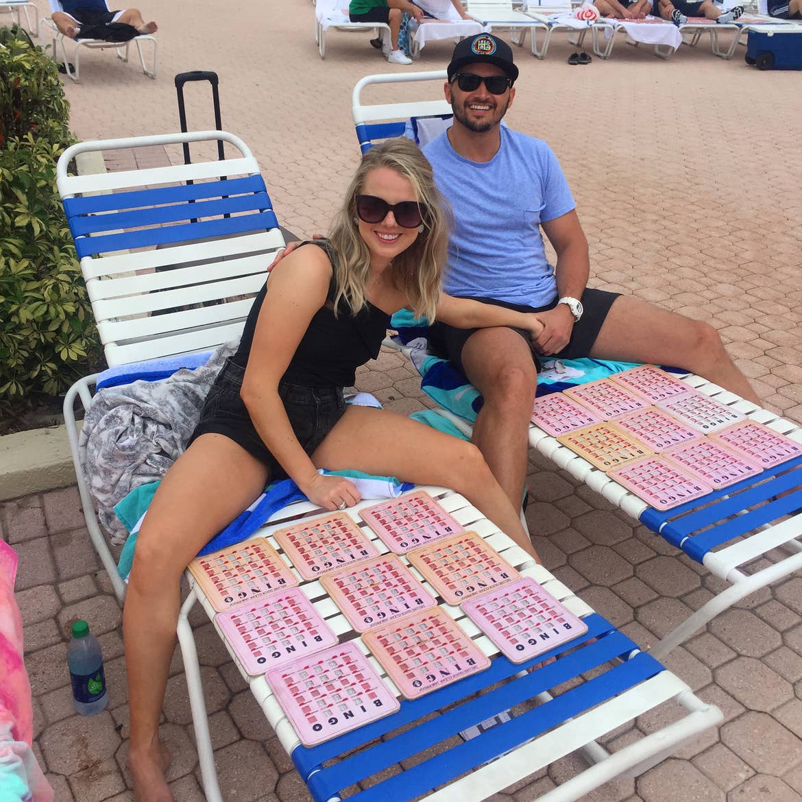 A caucasian woman wearing a black blouse and shorts (left) and Latinx male wearing a blue t-shirt, black shorts and a black hat (right) sit on two lounge chairs with numerous Bingo cards.