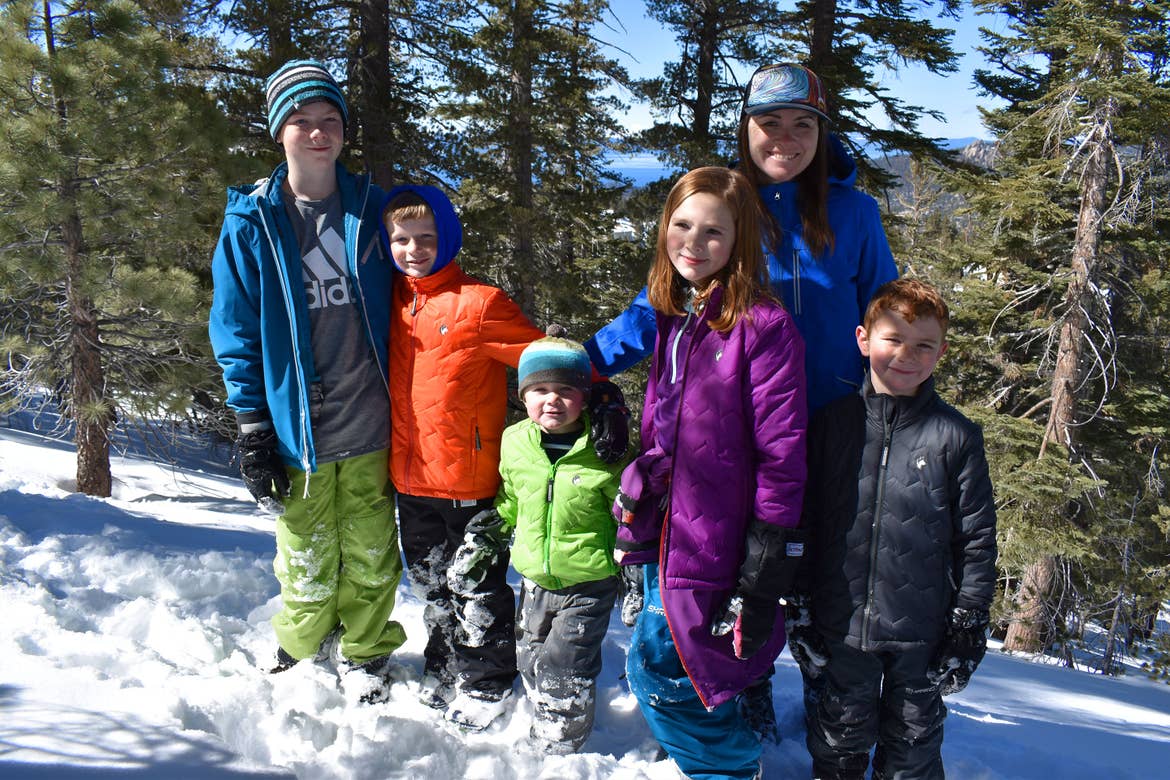A woman and five children wearing winter apparel stand on a snowy mountain surrounded by pine trees.