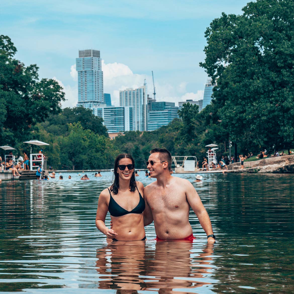 A woman in a black swimsuit and sunglasses stands with a man in swim trunks in the water as a skyline appears in the background.