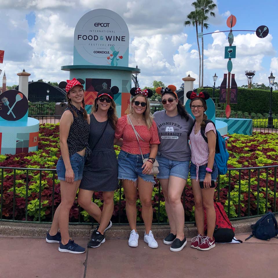 Five caucasian women wearing Mickey and Minnie ears stand in front of a display for the Epcot International Food & Wine Festival near the World Showcase Lagoon.