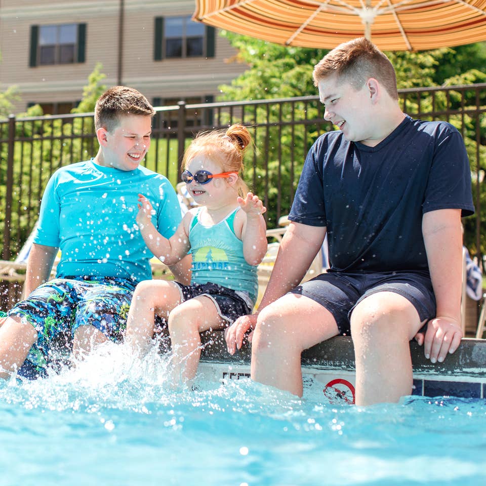 Two young caucasian tween boys (left and right) wearing swim shirts and shorts sit with their feet in a pool with a young caucasian girl (middle) wearing a swimsuit, swimming shorts and goggles as she splashes her feet.