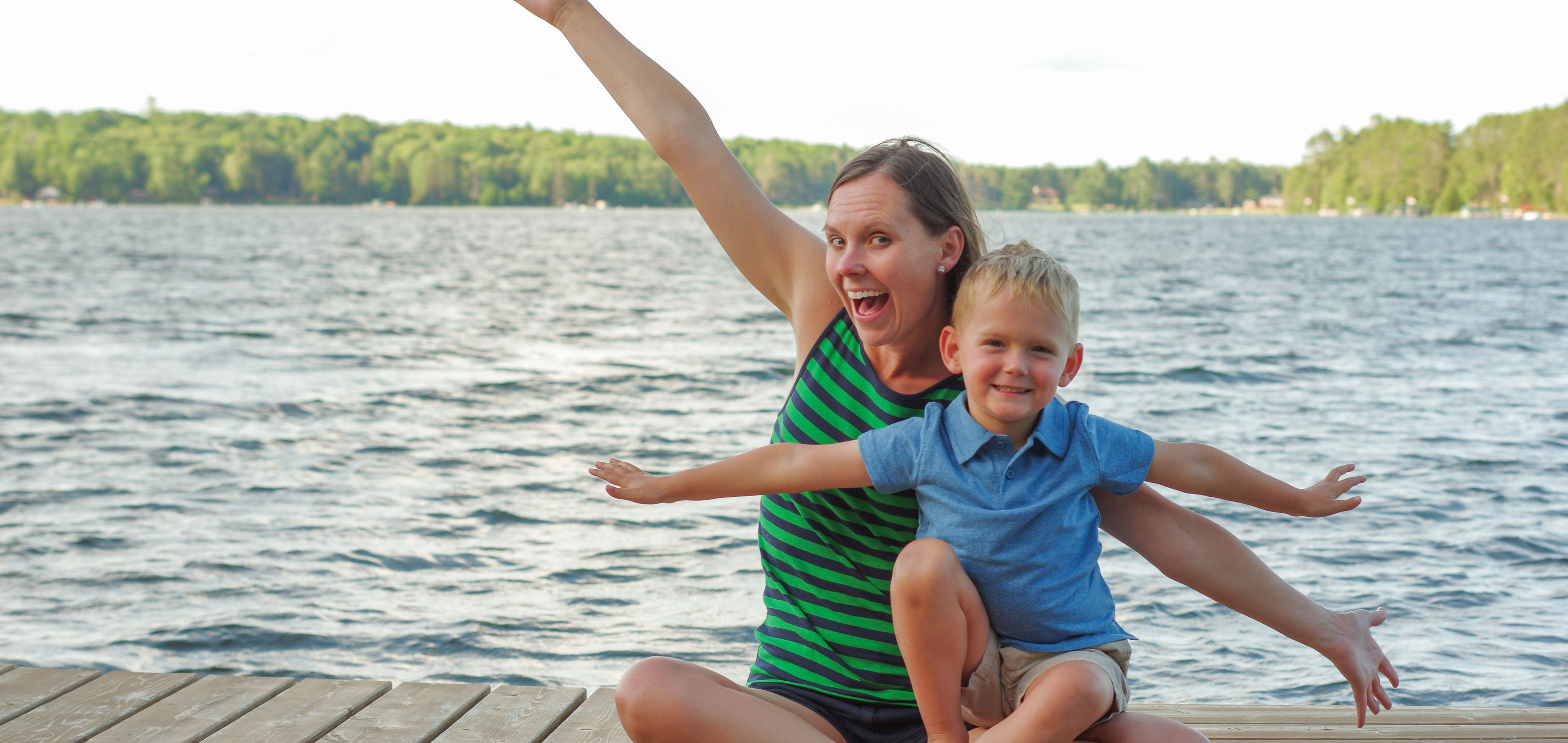 Author, Sarah Conroy, and son, Logan, sit on a lake pier mimicking an airplane with their arms.