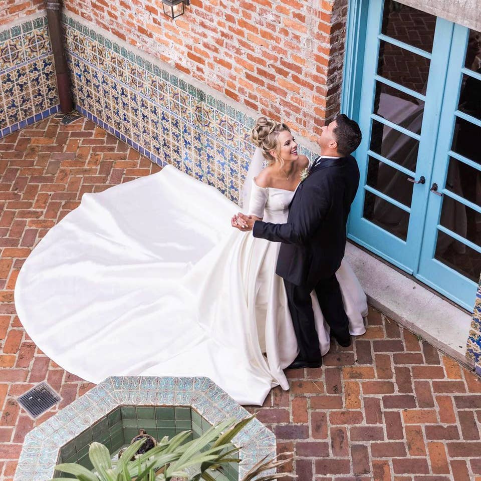 A Latinx groom wearing a black suit and (right) and a caucasian bride with a long train (left) dance with each other in front of a brick facade.