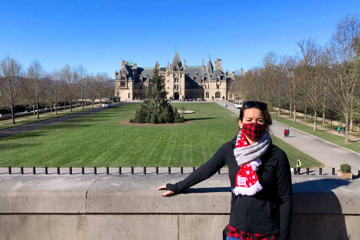 Featured Contributor, Jenn C. Harmon, stands in front of the Biltmore Estate wearing a buffalo plaid red and black face mask and festive scarf over a black jacket.
