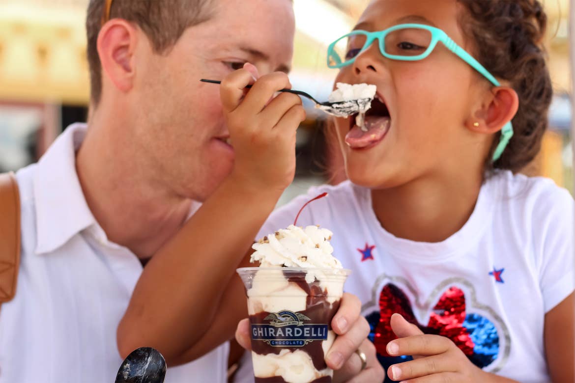 A Caucasian man wearing a white button-up shirt (left) and an Asian girl wearing a sparkly, white Minnie Mouse t-shirt and blue glasses (right) share an ice cream sundae from Ghirardelli Chocolate outdoors.