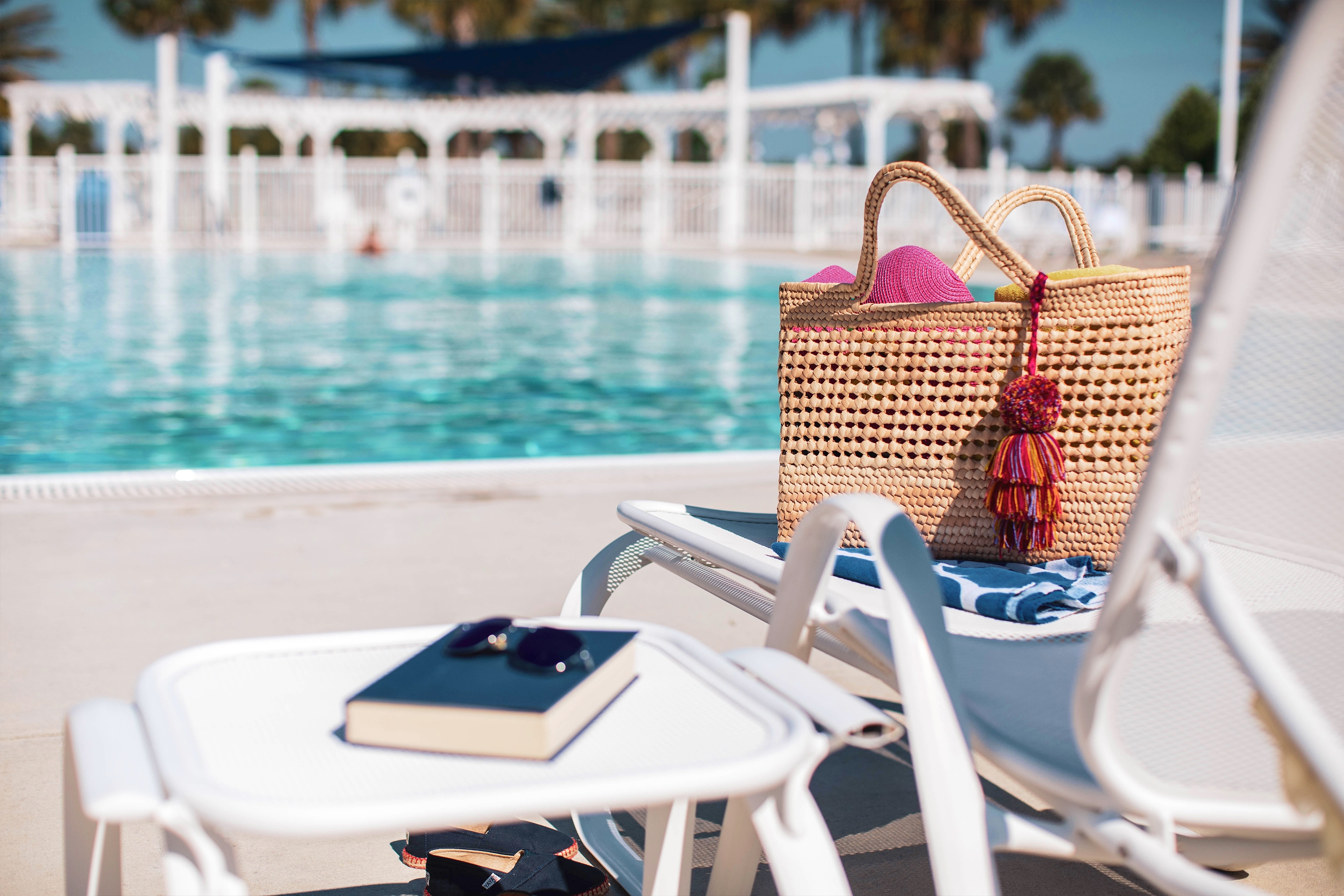 A pool chair lined up at edge of outdoor pool at Orlando Breeze Resort in Florida has a beach bag, towels and a book and sunglasses placed on the chair and table.