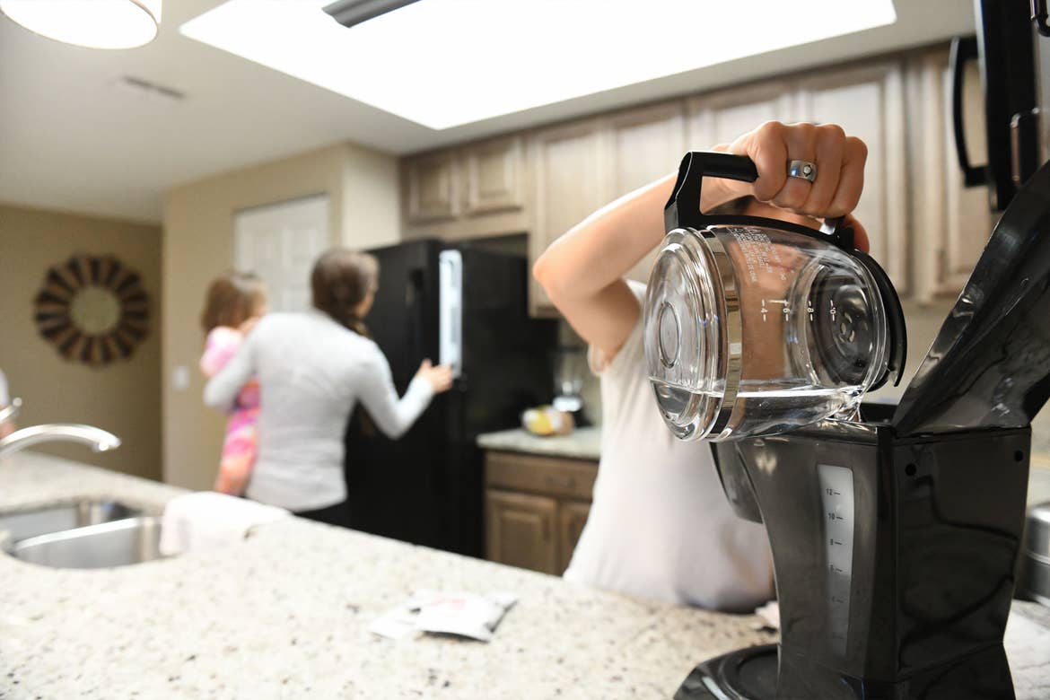 From left to right: A woman holding a girl toddler opens a black refrigerator as another woman fills a coffee maker.
