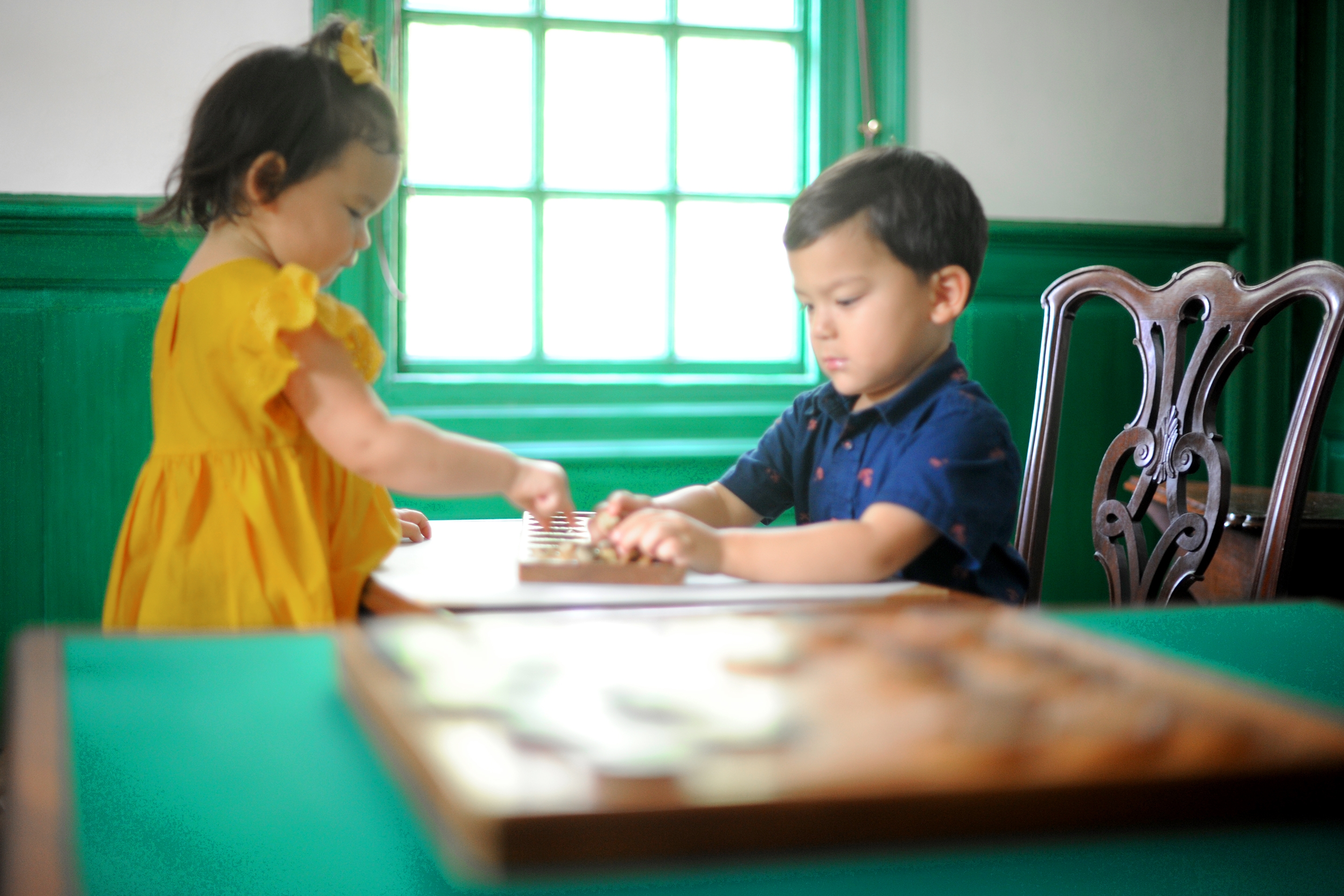 Two Asian Pacific Islander toddlers (left to right: A girl and a boy) sit near a table playing Mancala in a green and white room.
