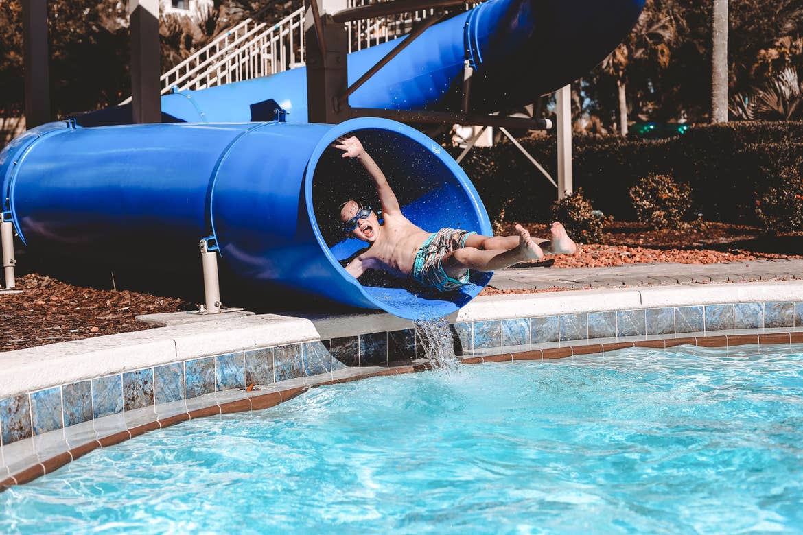 Grey wears swim trunks and goggles as he emerges from a blue water slide at River Island.