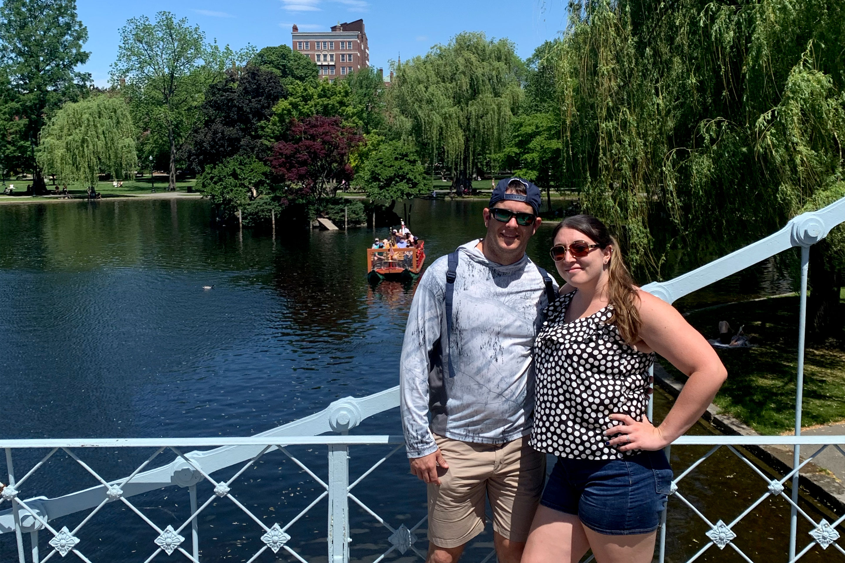 A man (left) and woman (right) stand on a bridge over water with willow trees and a swan boat floating in the back.