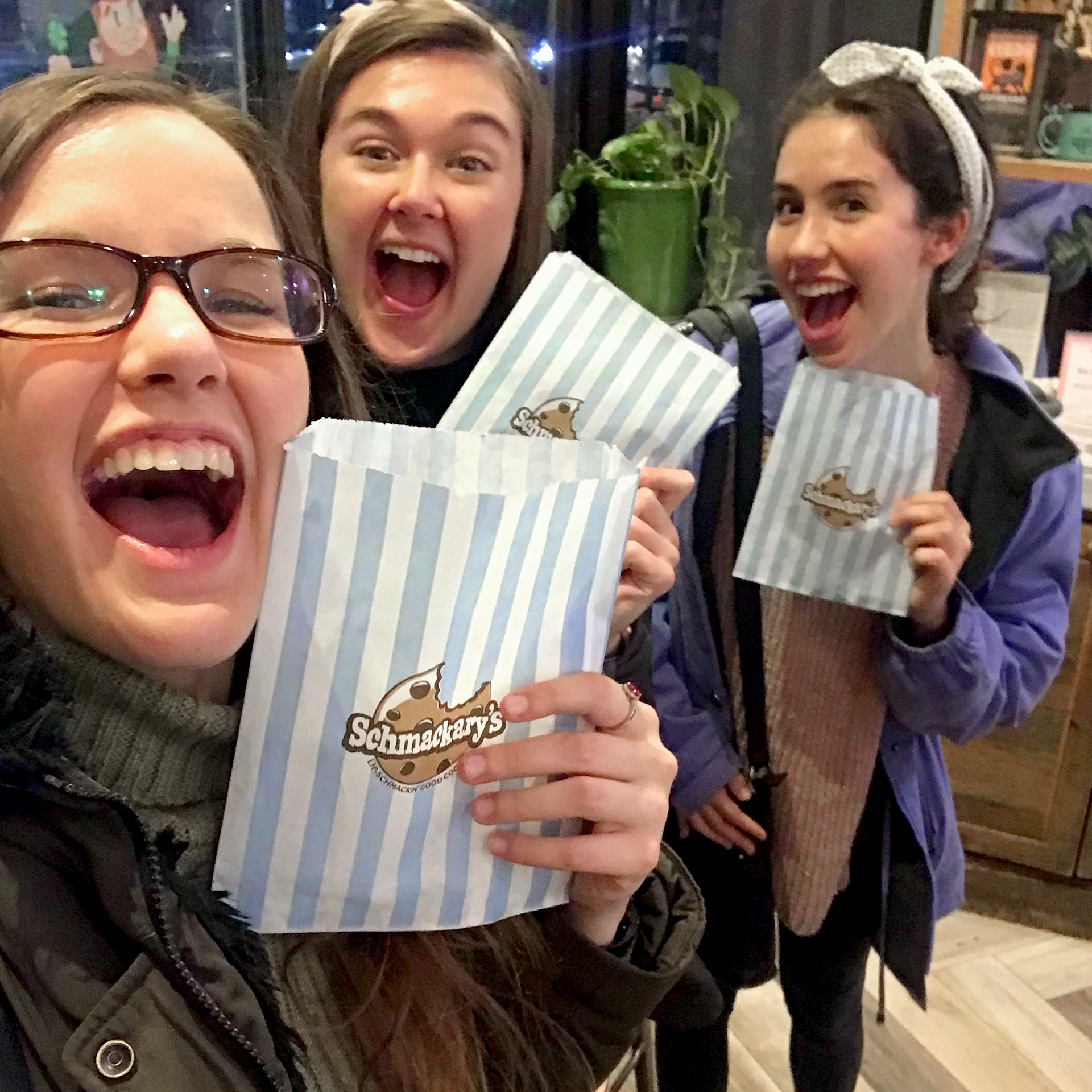 Three caucasian women in winter jackets hold a wax paper with blue and white stripes from 'Schmackary’s ' in New York City.
