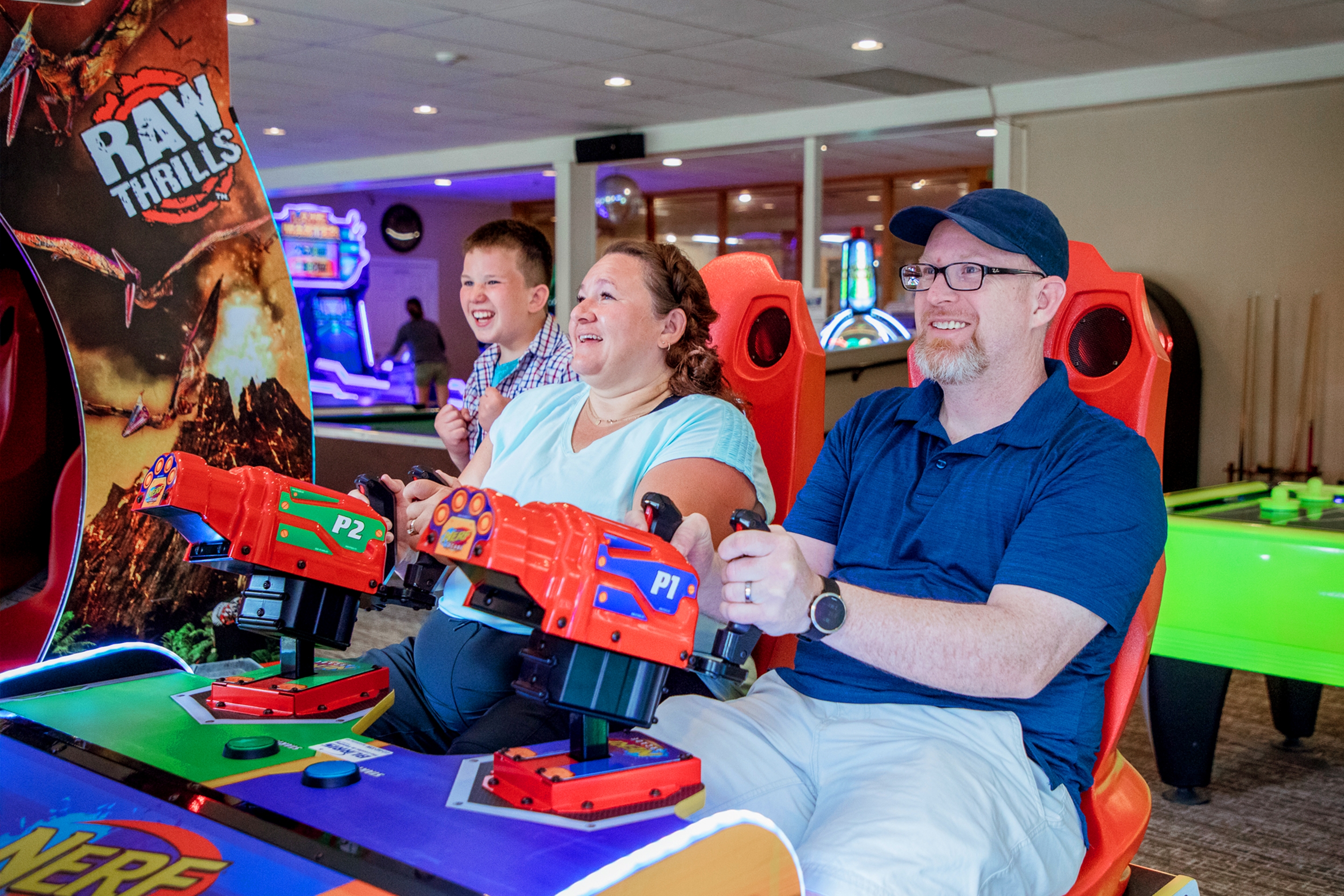 A caucasian man (right) wearing a navy baseball cap and polo is seated next to a caucasian woman (left) wearing a light blue t-shirt in an arcade gaming seat behind shooters as a tween boy (back-left) watches from behind the seat.