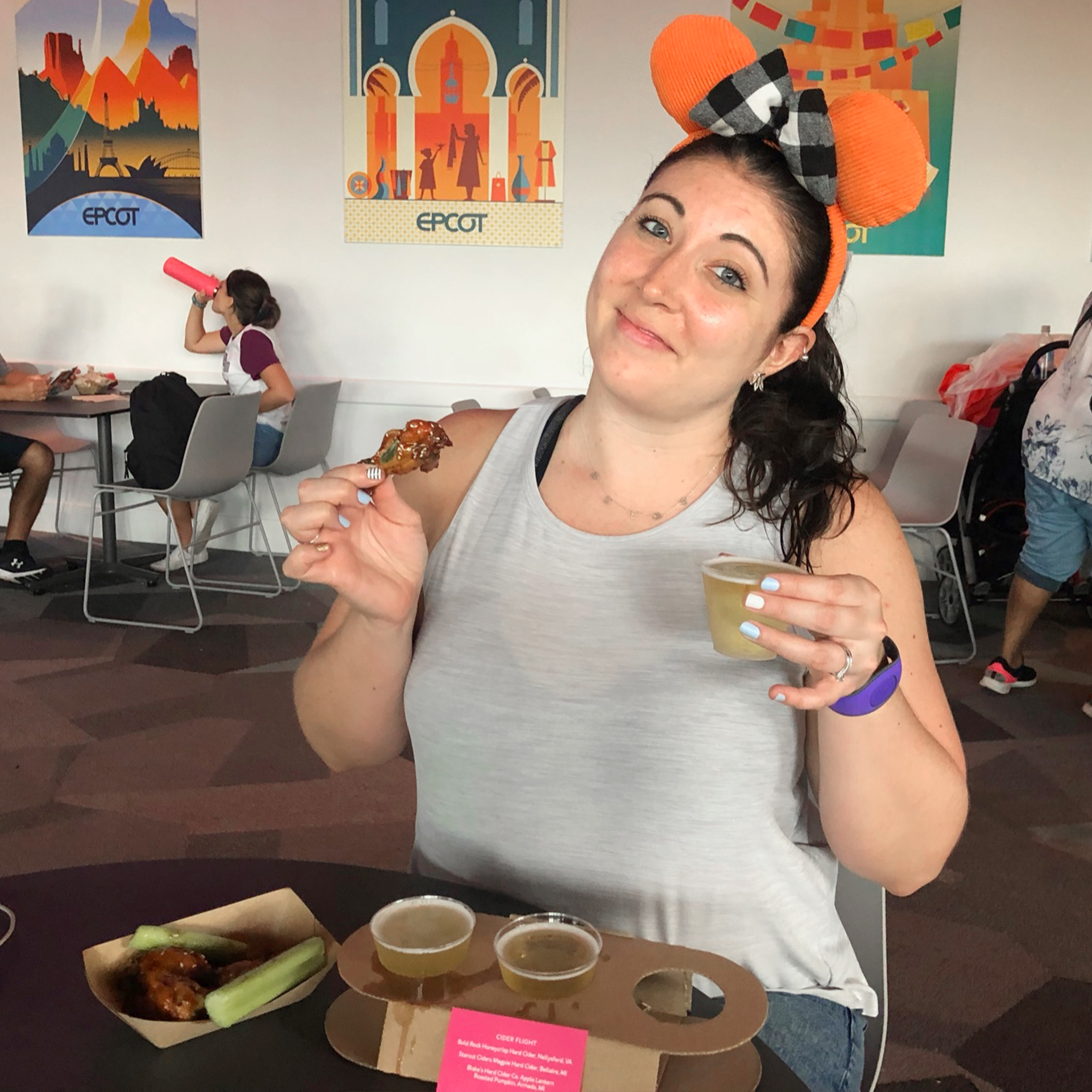 A caucasian woman wearing orange Mickey ears and a grey tank top holds a chicken thigh in her right hand, and cider in her left hand indoors.