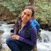 Featured author, Andrea Beltran, poses near a creek wearing a blue hiking backpack and purple pull-up sweater.
