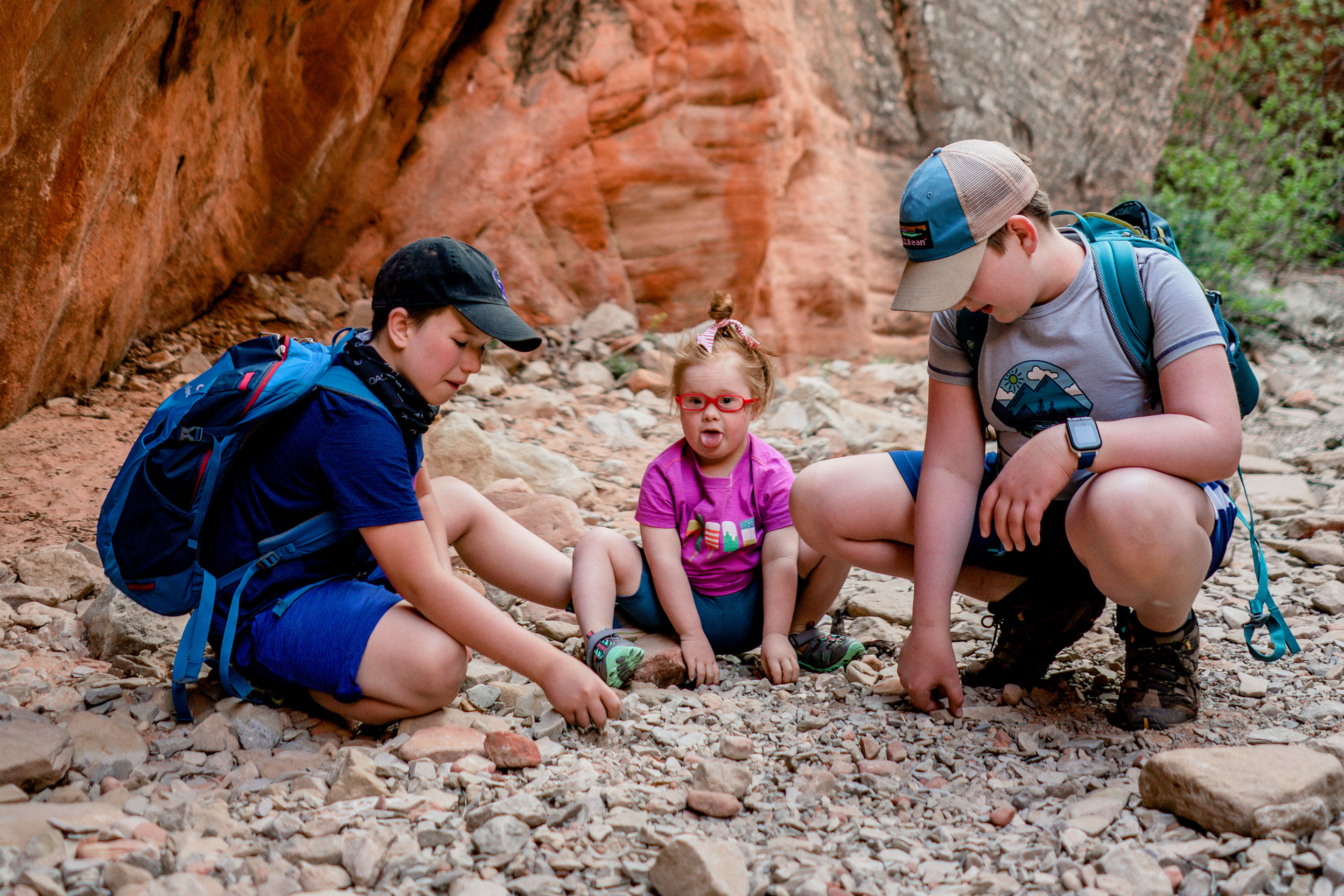 Featured Contributor, Melody Forsyth's two sons (left and right) sit with daughter, Ruby (middle) in front of a rock formation touching stones while wearing hiking backpacks.