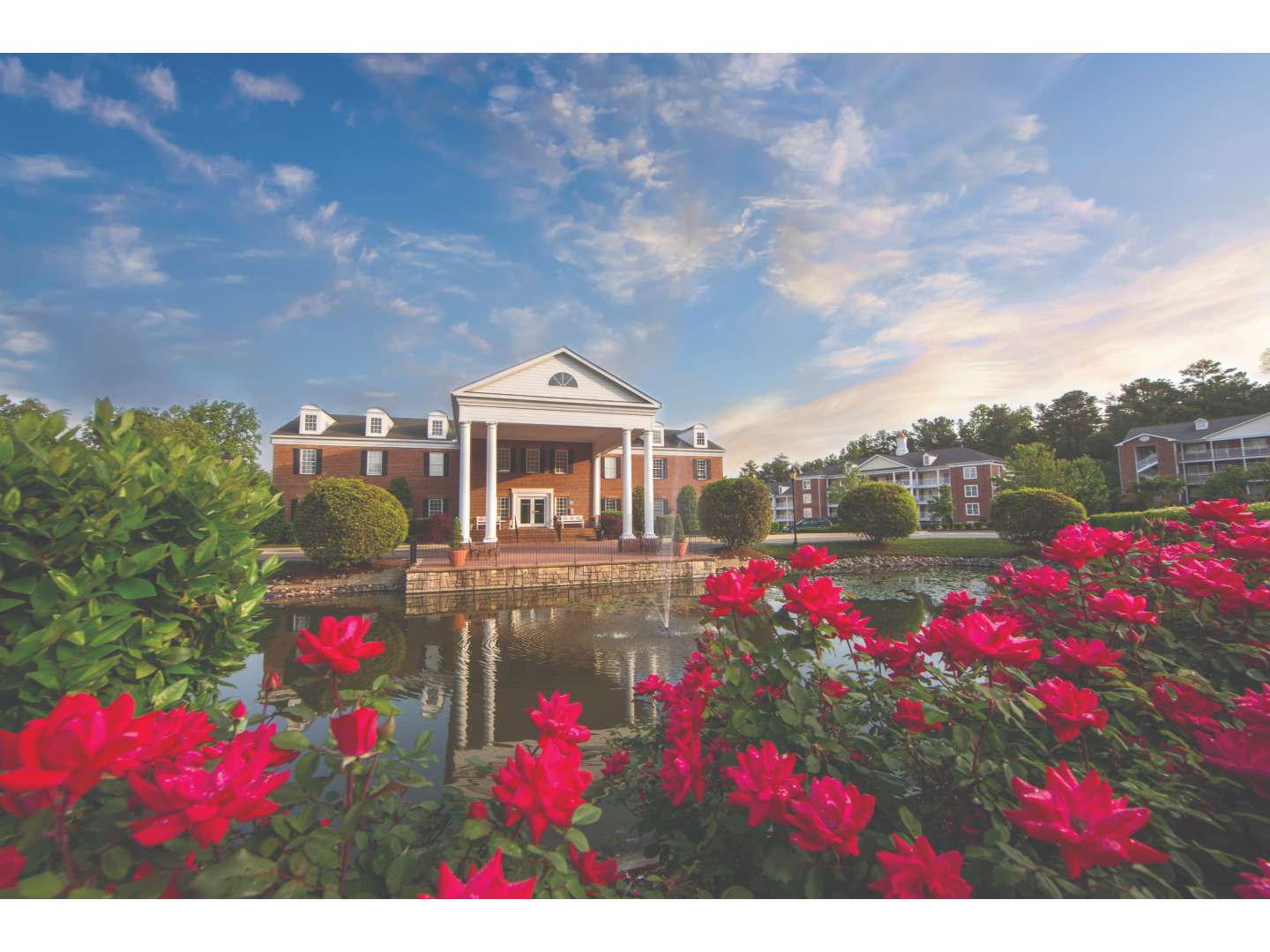 Large brick mansion with 4 white columns, a fountain, and bright flowers.