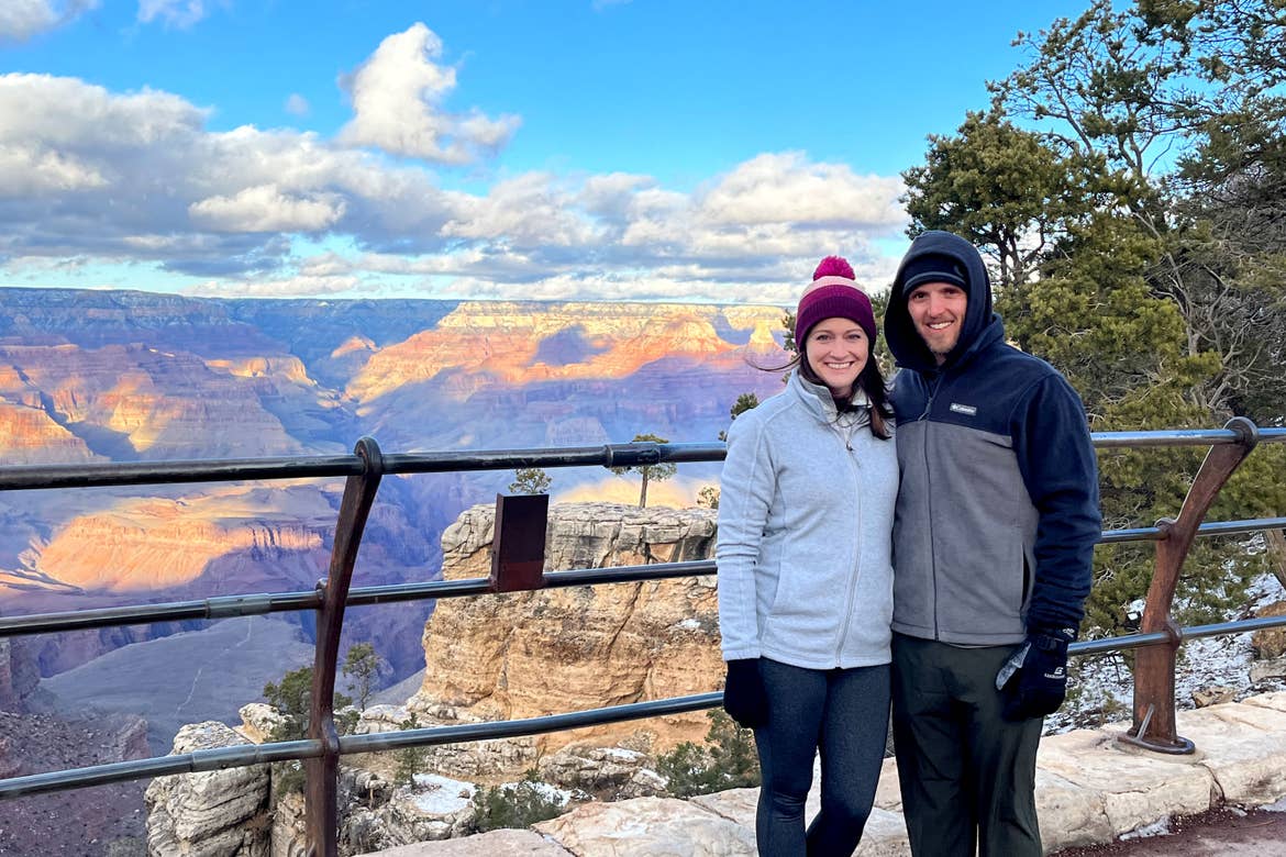 A woman wearing a white fleece jacket and magenta knitted cap stands next to a man in a blue-grey fleece jacket with a black knitted cap on a hiking trail near a rail-guard overlooking the Grand Canyon.