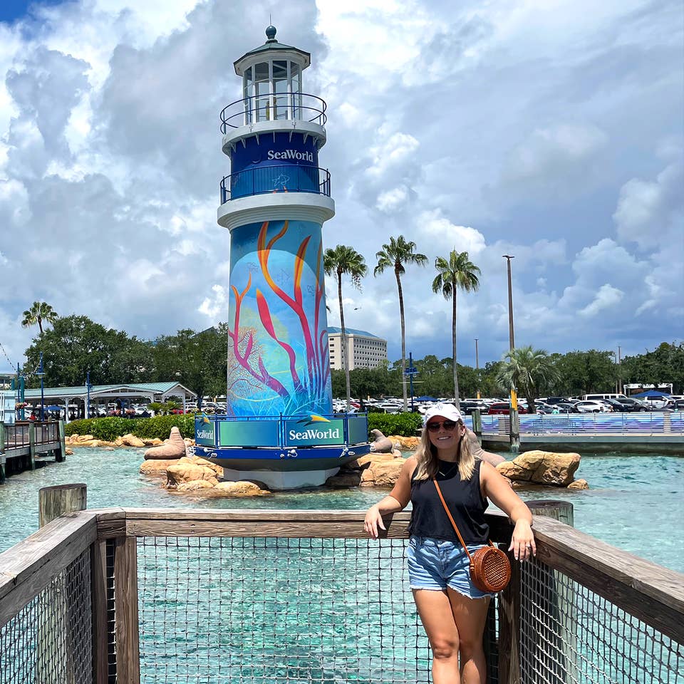 A Caucasian female wearing a pink baseball cap and black tank top stands near the lighthouse in SeaWorld Orlando.