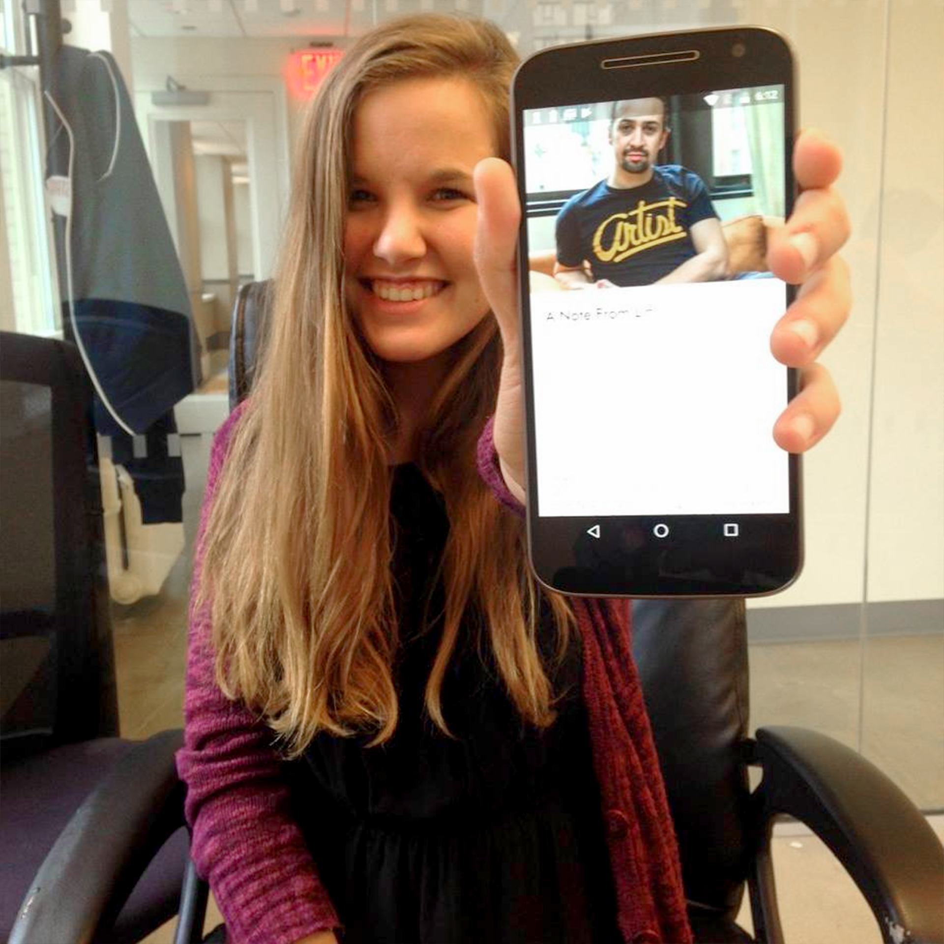 A caucasian woman with long ash-blonde hair wears a red cardigan while holding a mobile device with a picture of Lin Manuel-Miranda for the 'Hamilton' Ticket Lottery.