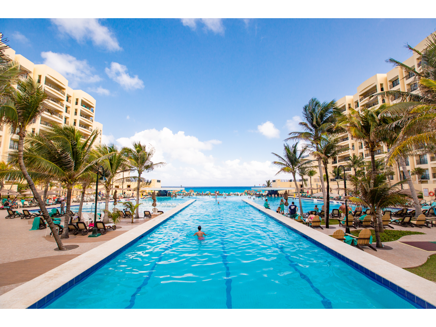 Long pool surrounded by resort buildings and palm trees, with the ocean behind.