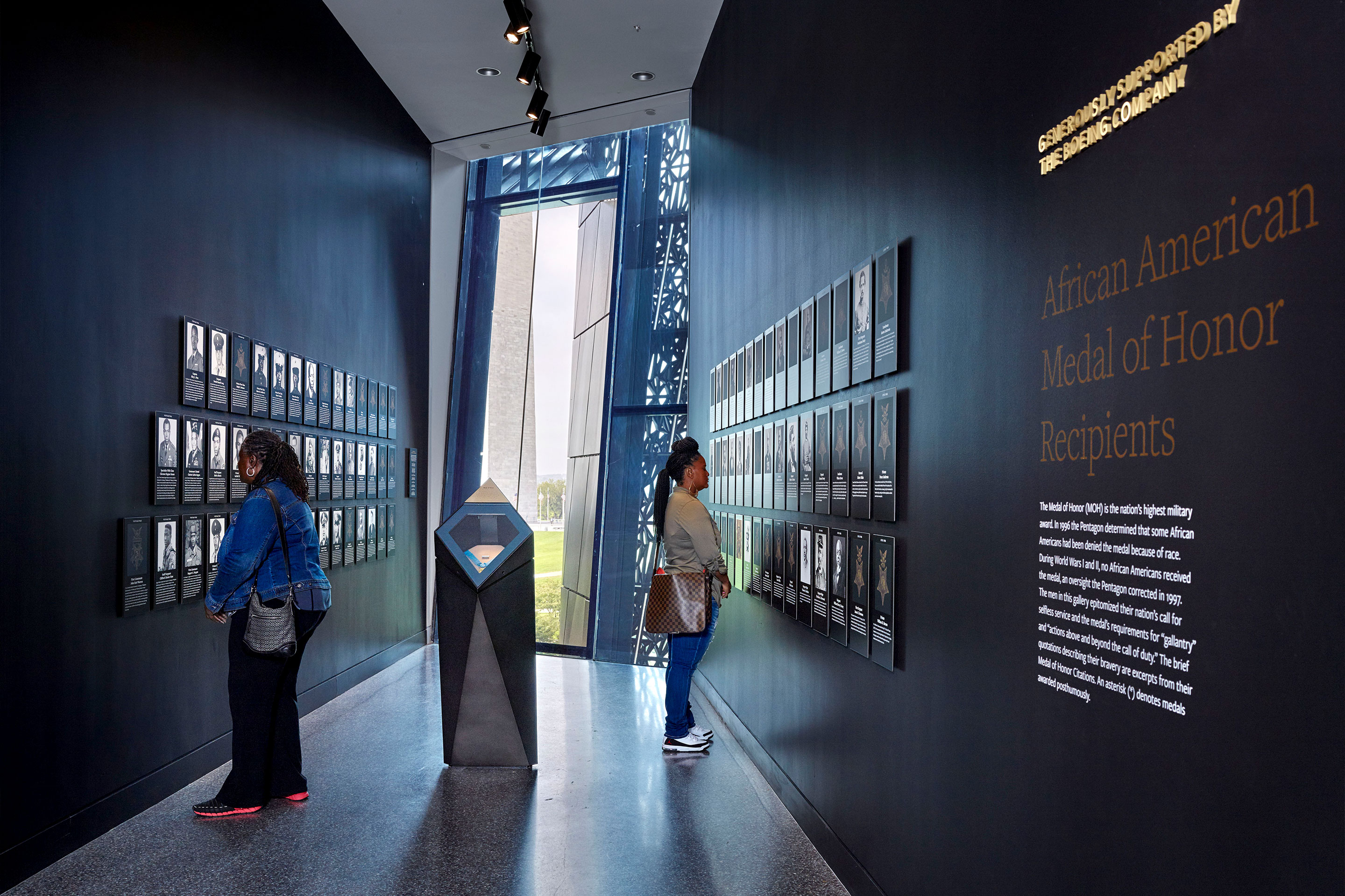 Two women stand in a black hallway curated with photography, statues and supportive signage.