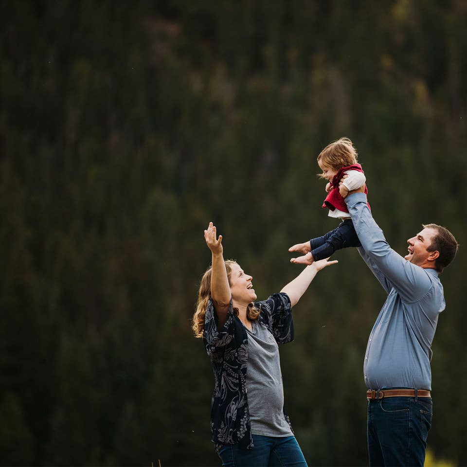 A mother (left) lifts her expressive arms as a father (right) holds up their child in front of a wooded area.