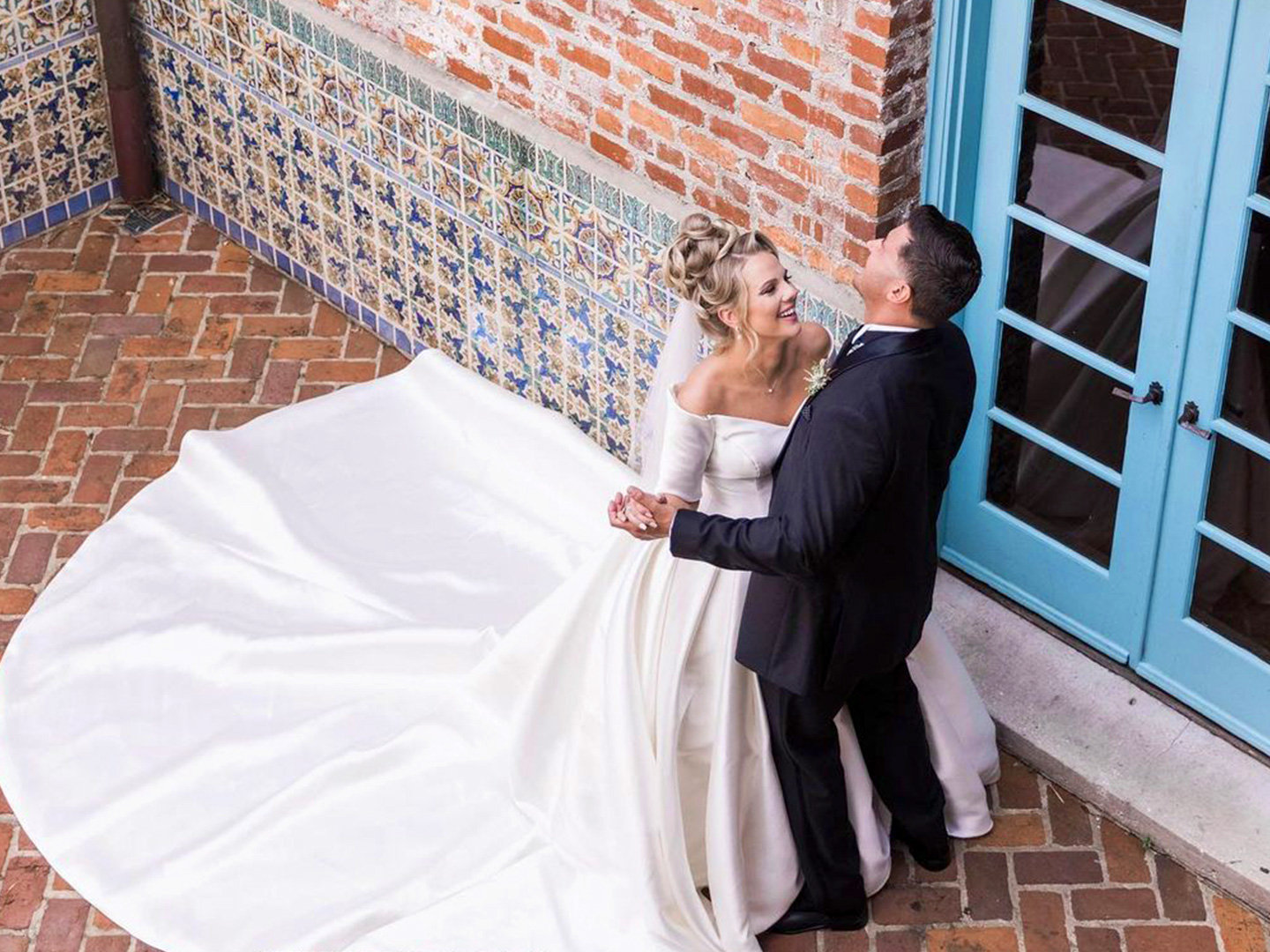 A Latinx groom wearing a black suit and (right) and a caucasian bride with a long train (left) dance with each other in front of a brick facade.