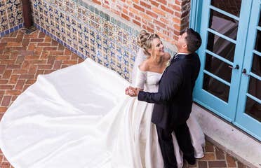 A Latinx groom wearing a black suit and (right) and a caucasian bride with a long train (left) dance with each other in front of a brick facade.