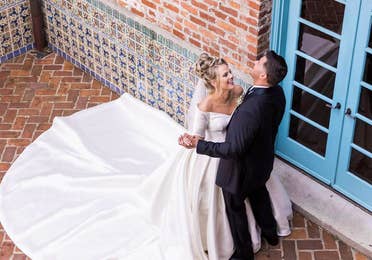 A Latinx groom wearing a black suit and (right) and a caucasian bride with a long train (left) dance with each other in front of a brick facade.