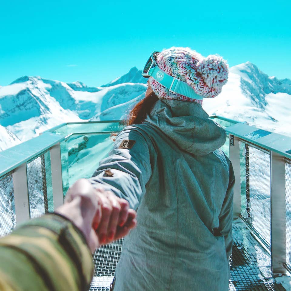 A woman holds a man's hand while wearing winter apparel and ski goggles looking off at snowcapped mountains under a blue sky.