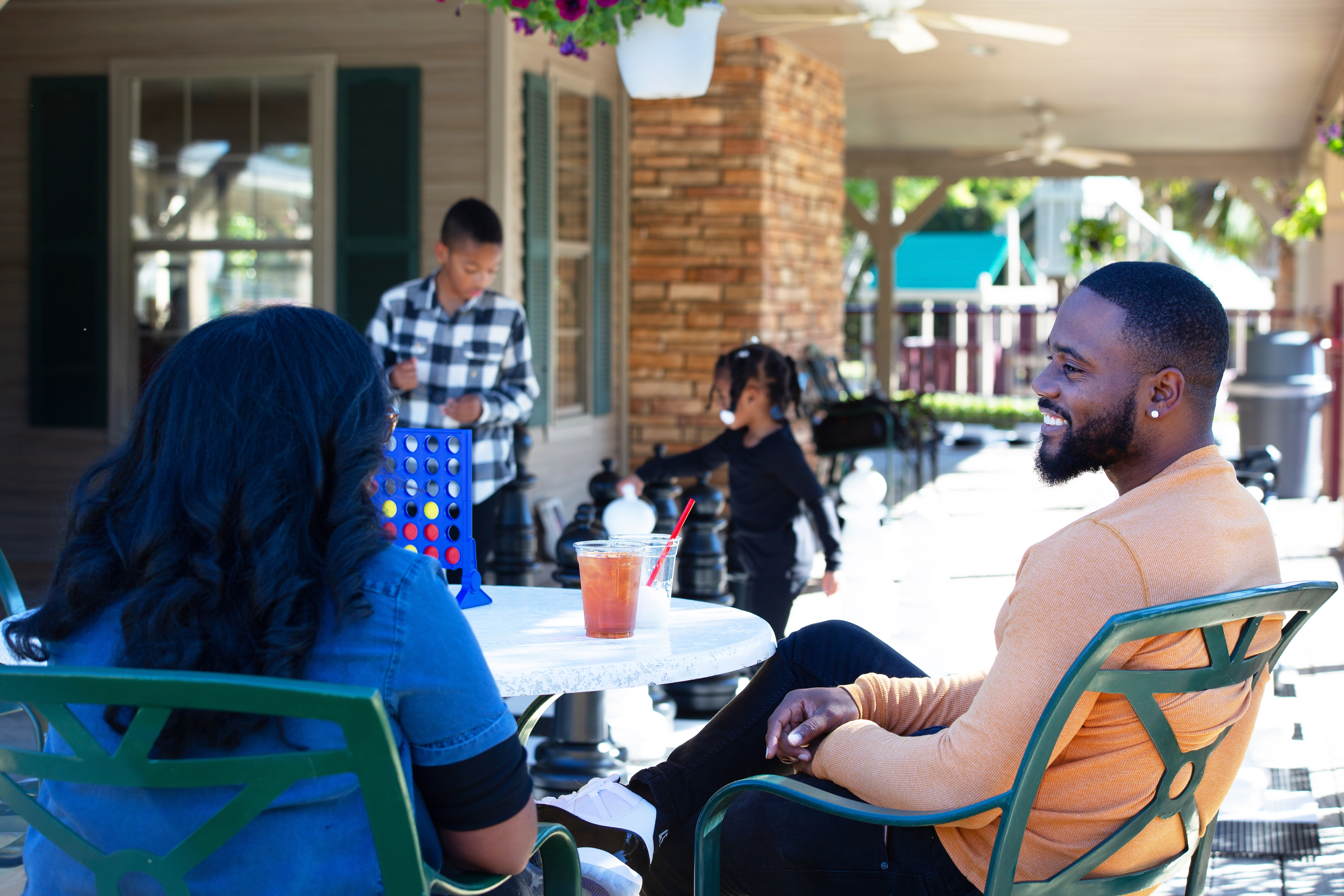 Tina and her husband relaxing at an outdoor table with drinks while the kids play with giant chess pieces. 