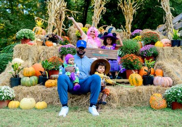 A man, two young toddlers, and two young girls sit on a festive stack of hay bails.