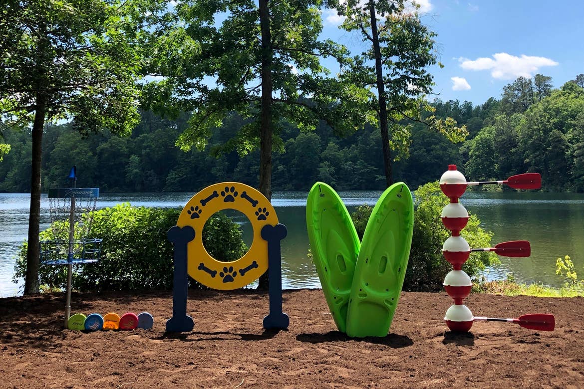 Various colored recreational items, such as a Disc Golf post, a Dog obstacle ring, two kayaks, and buoys, form the word, 'LOVE' in an outdoor park near a lake.