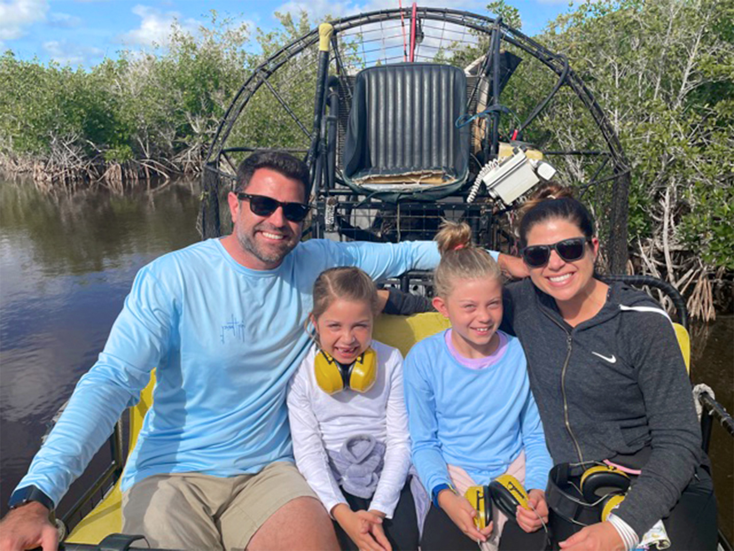 A caucasian woman (left) wearing sunglasses and a black zip-up hoodie, two Caucasian girls (middle) and a caucasian male (right) wearing sunglasses and a blue long-sleeve shirt sit on an airboat in the Everglades.