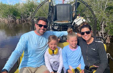 A caucasian woman (left) wearing sunglasses and a black zip-up hoodie, two caucasian girls (middle) and a caucasian male (right) wearing sunglasses and a blue long-sleeve shirt sit on an airboat in the Everglades.