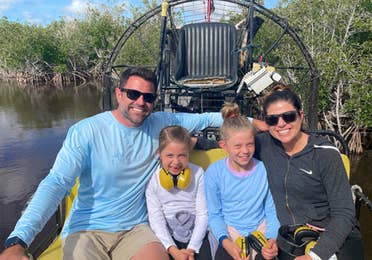 A caucasian woman (left) wearing sunglasses and a black zip-up hoodie, two caucasian girls (middle) and a caucasian male (right) wearing sunglasses and a blue long-sleeve shirt sit on an airboat in the Everglades.