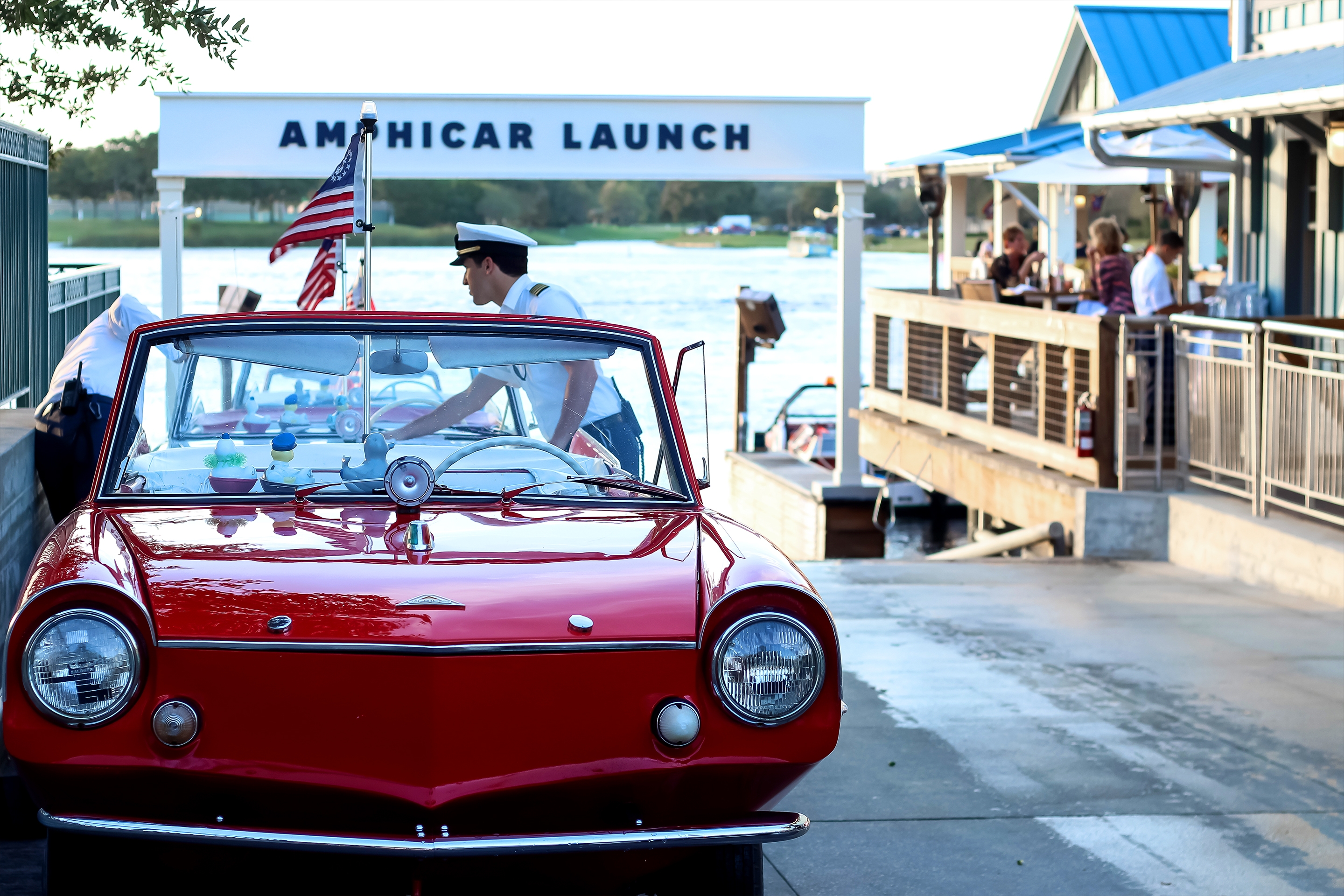 A Caucasian man wearing Nautical Captain attire preps a red, amphibious car on the launch pad near a white sign that reads, ‘Amphicar Launch.’