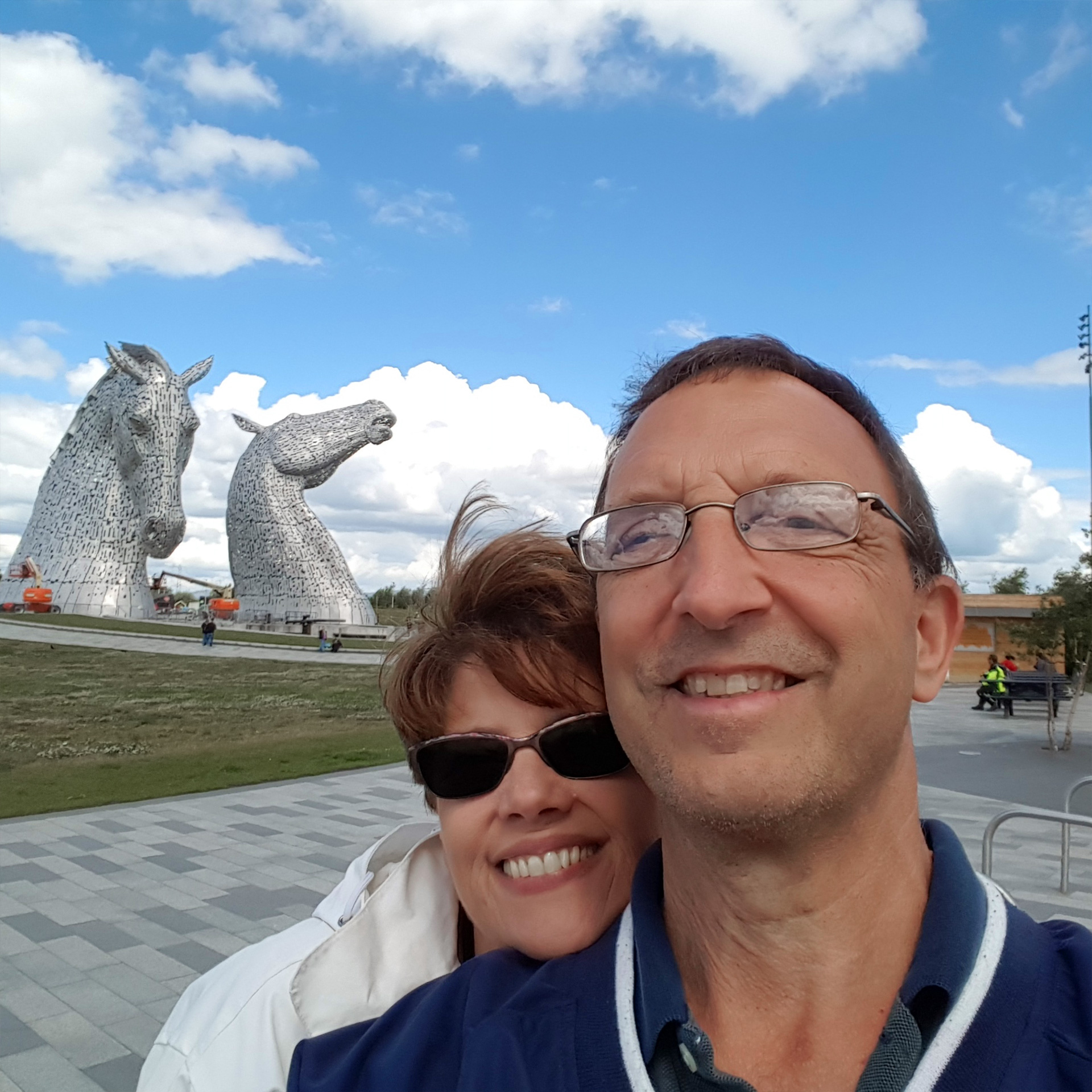 A caucasian man (right) wearing a navy windbreaker and a caucasian woman (left) standing behind him in a white windbreaker jacket stand in front of the Kelpies in Falkirk, Scotland.