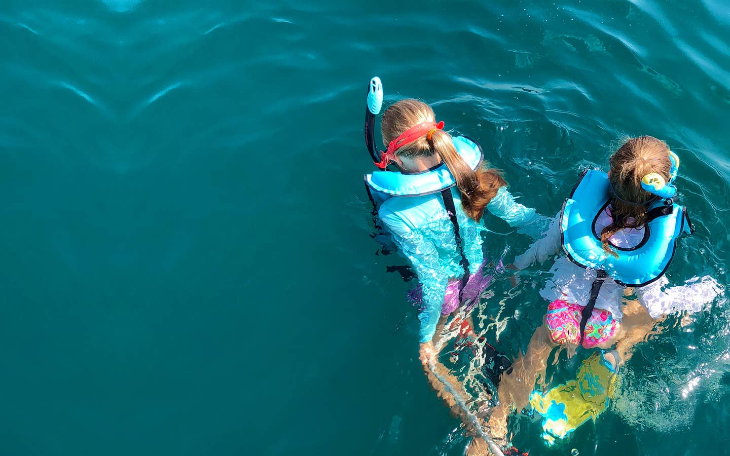 Featured Contributor, Chris Johnston's two daughters, Kyler (right) and Kyndall (left), wear multi-colored snorkel gear while holding onto a rope in the ocean.