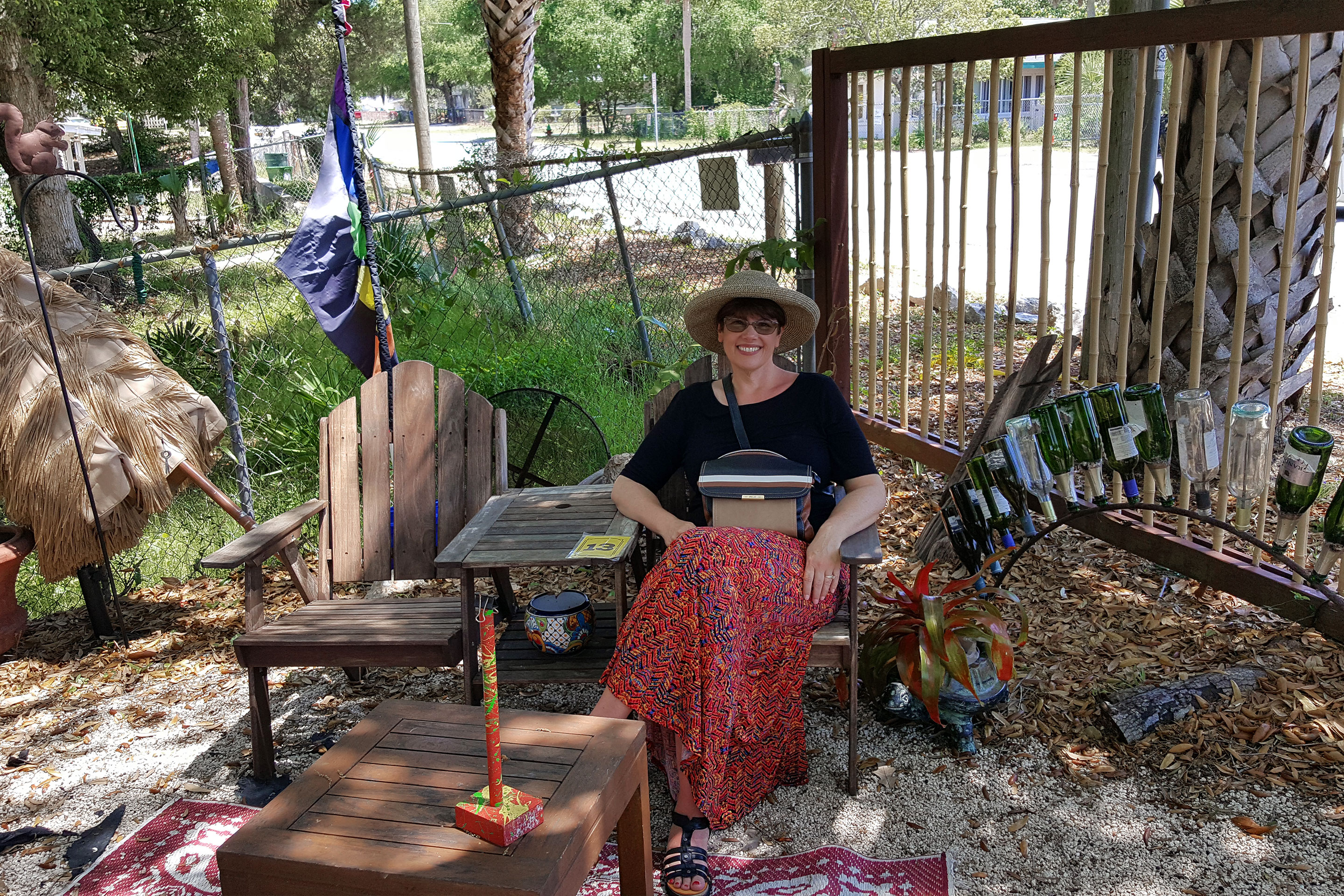 A woman in a sunhat, black shirt, and floral, red skirt sits on a wooden chair surrounded by glass art sculptures outdoors.