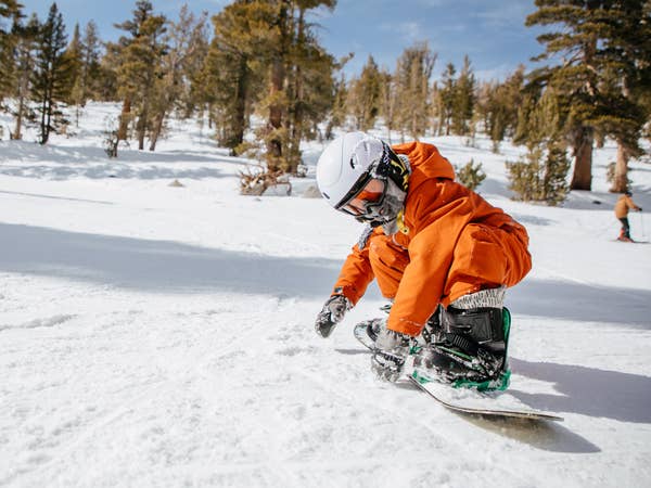 A boy wearing orange snow jacket, pants and white helmet makes his way down a snowy slopes on a snowboard.