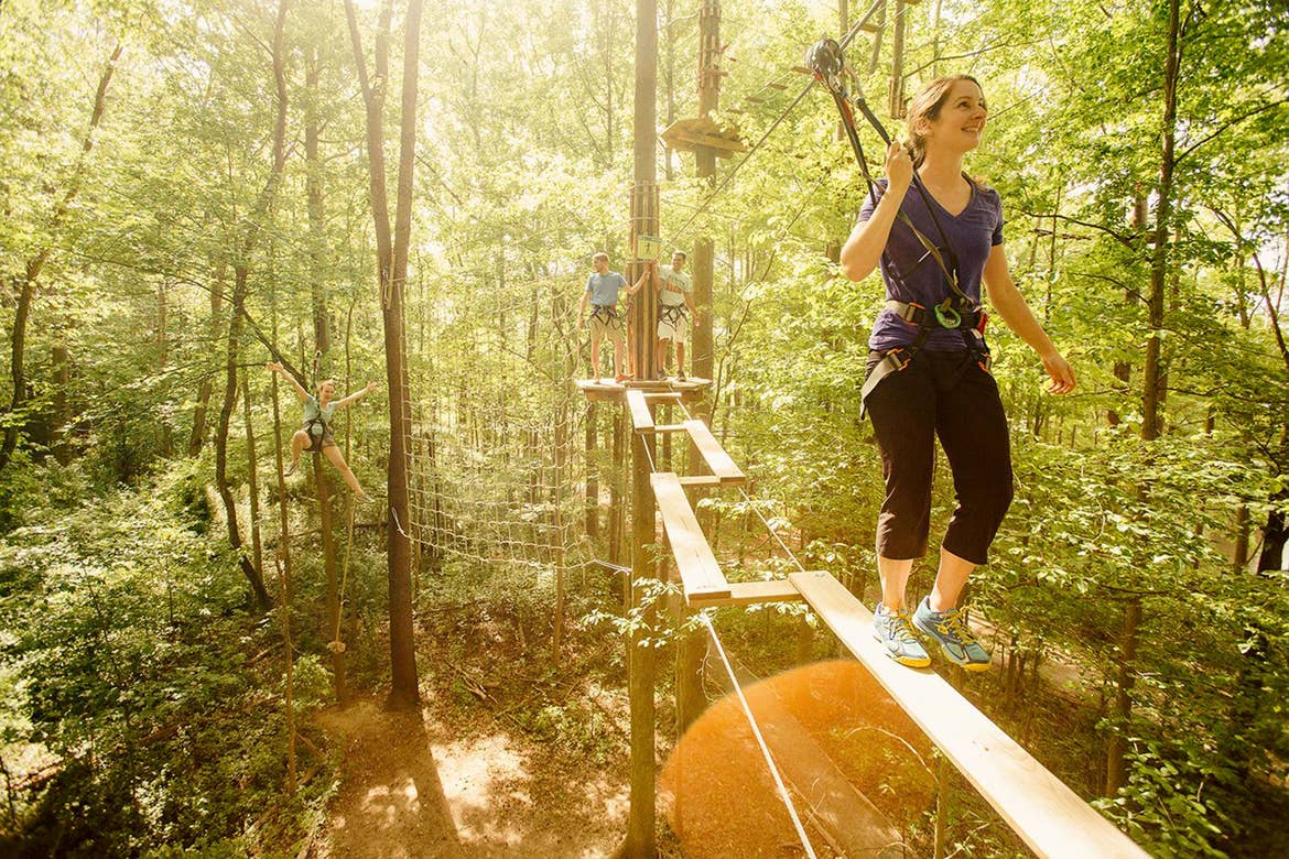 A woman in a black shirt, pants, and sneakers has a safety harness across a high ropes course in the woods.