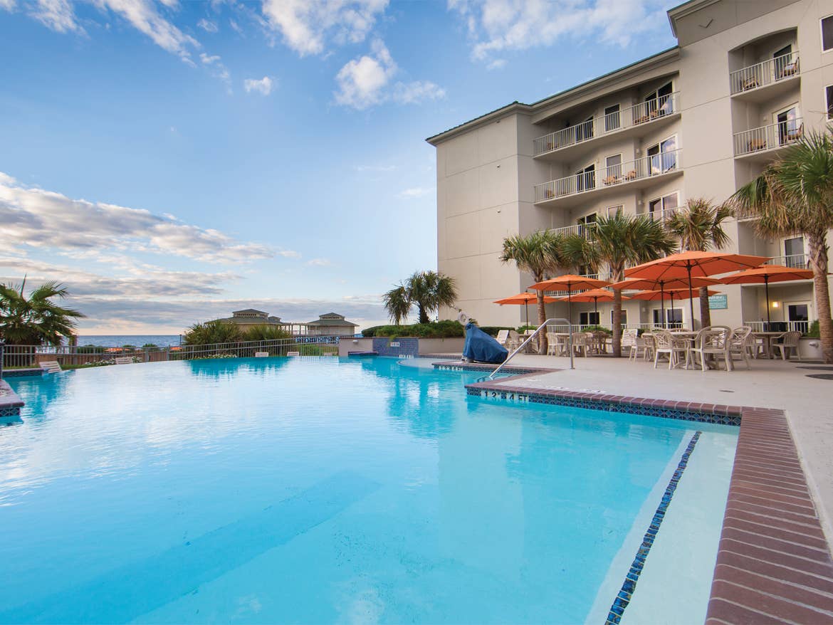 Large blue pool with orange umbrells and tables and beach in distance.