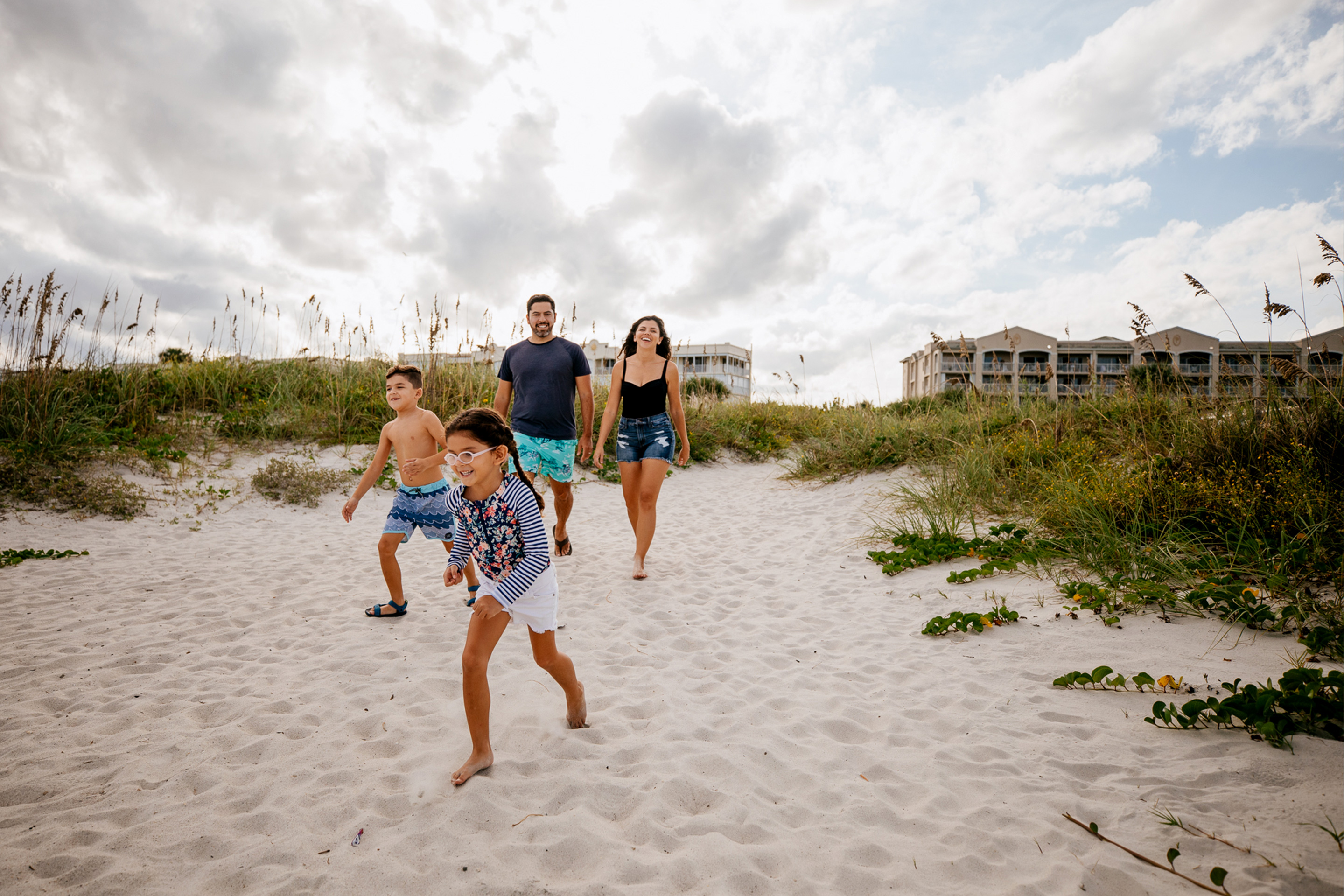 A young boy and girl run on sand in front of a man and woman wearing swimwear in front of a beachfront resort.