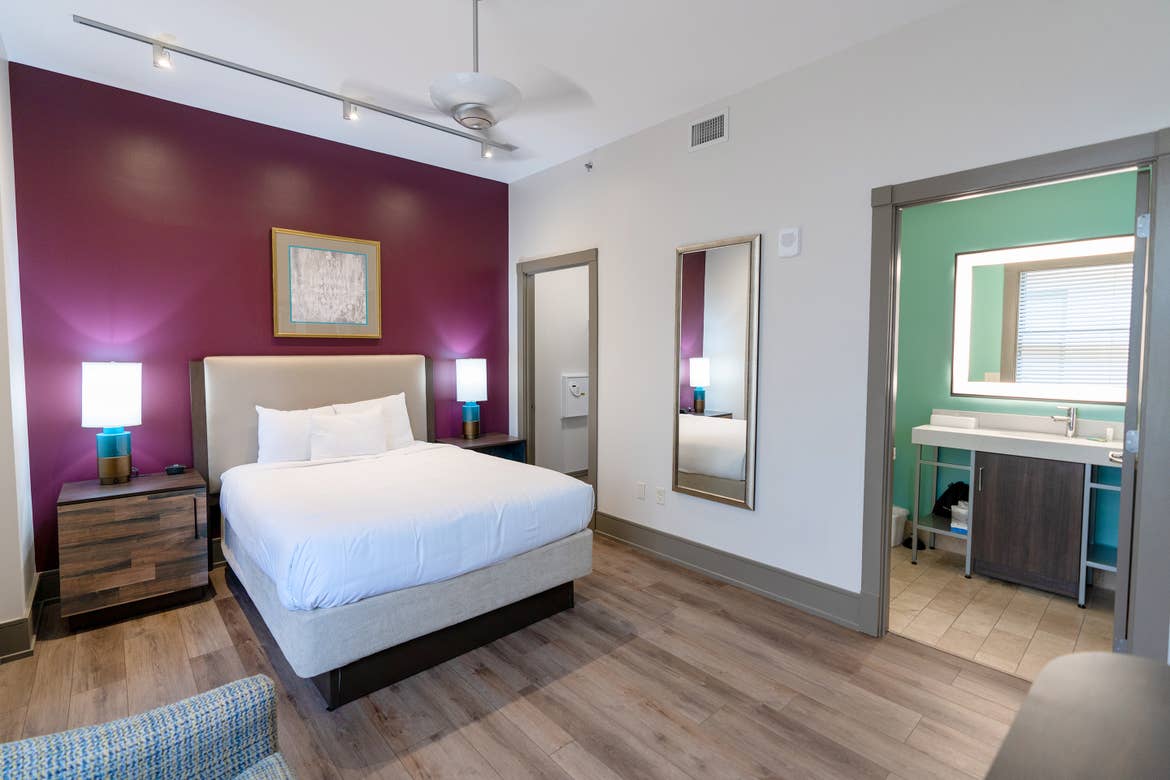 The master bedroom of our one-bedroom standard villa at our New Orleans resort furnished with a queen-sized bed with white sheets, two lamps and nightstands, a purple feature wall and a master bathroom to the right with green walls.