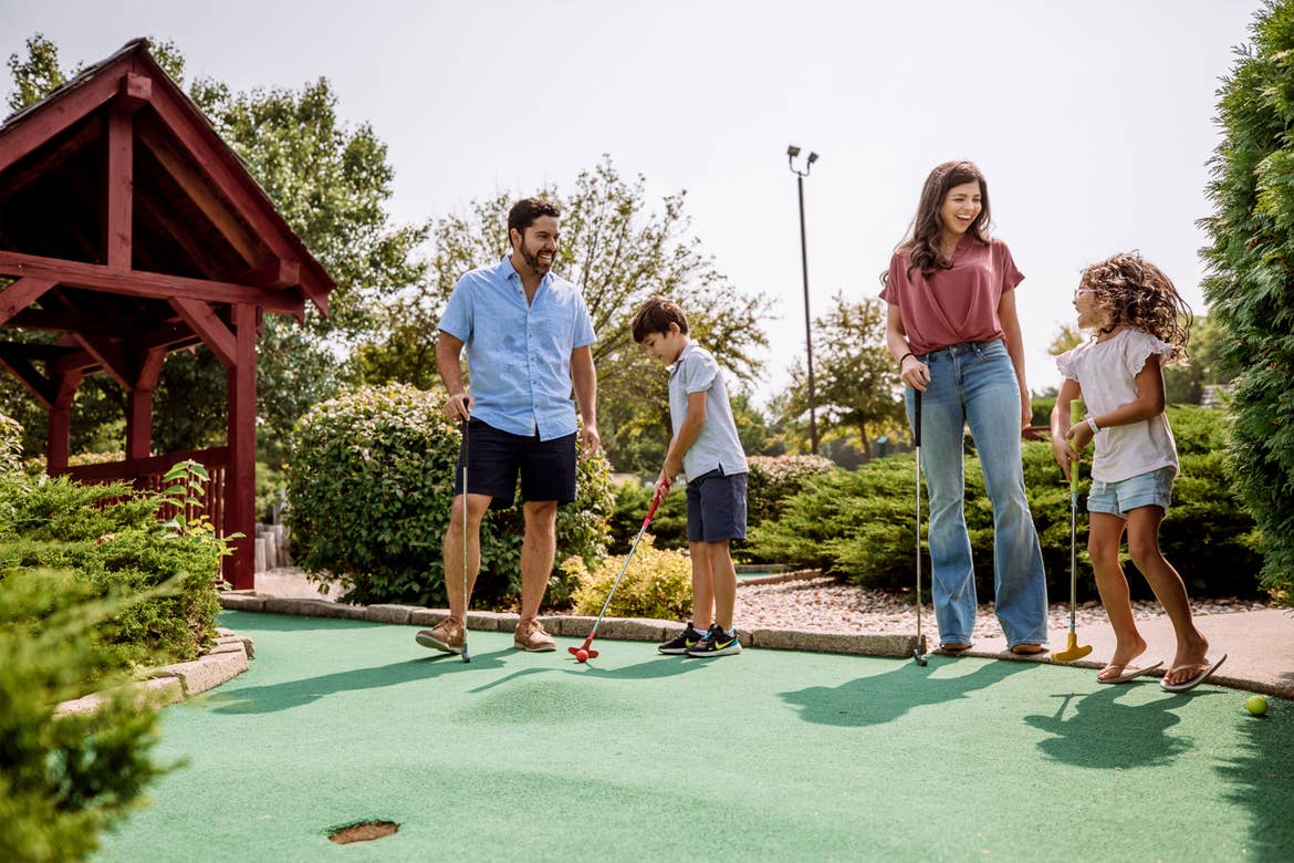 A family of four wear denim and play on an outdoor mini-golf course.