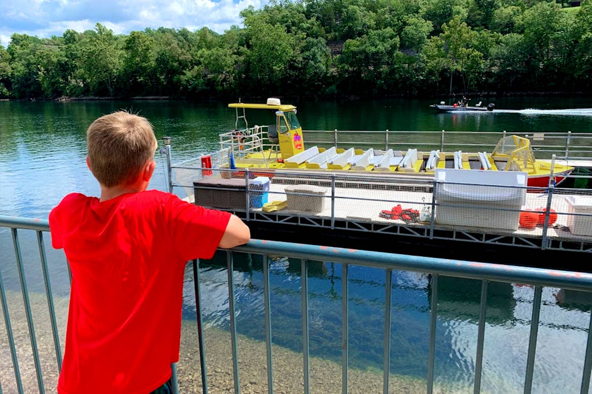 A boy in a red shirt sits on a dock overlooking the river where a yellow Jet Boat is docked.