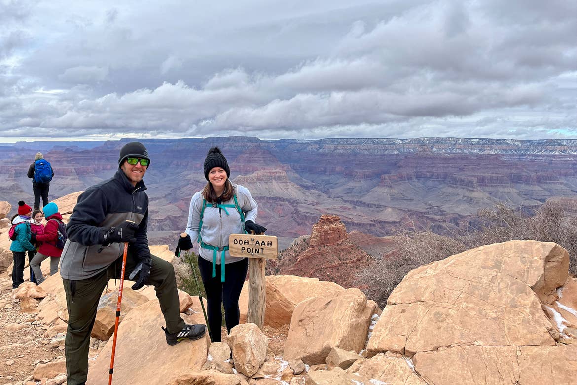 A woman wearing a white fleece jacket and magenta knitted cap stands next to a man in a blue-grey fleece jacket with a black knitted cap on a hiking trail near a sign that reads, ''OOH AHH Point' overlooking the Grand Canyon.